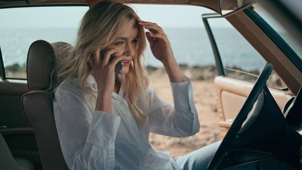 Γυναίκα οδηγός μιλάει τηλέφωνο κάθεται σε ρετρό αυτοκίνητο στην ηλιόλουστη ακτή του ωκεανού κοντά. Ελκυστική ευτυχής κορίτσι ταξιδιώτη απολαμβάνουν τηλεφωνική κλήση κάθεται στο αυτόματο τιμόνι. Υπέροχη ξανθιά μιλώντας smartphone. - Φωτογραφία, εικόνα
