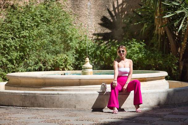 South American woman, young, beautiful, blonde with white top, pink pants and sunglasses, sitting in a nice fountain, sunbathing, relaxed and calm. Concept travel, destinations, Europe, vacation. - Photo, Image