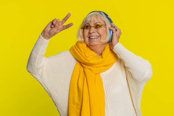 Opgewonden senior stijlvolle oude vrouw luisteren naar muziek via de hoofdtelefoon, dansen disco partij gekloot rond het hebben van plezier expressieve gebarentaal handen. Gelukkig oudere oma gepensioneerde op gele achtergrond - Foto, afbeelding