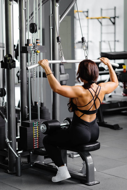 Muscular atlético fisiculturista feminino em terno preto puxado em simulador de esportes no ginásio. Os músculos das costas estão muito tensos. - Foto, Imagem