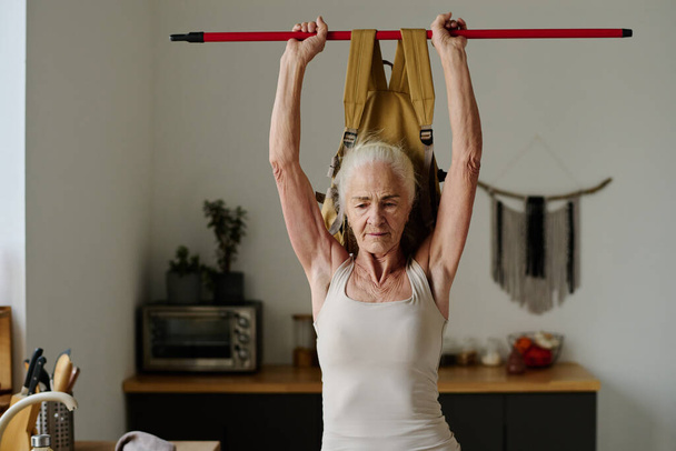 Senior vrouw met wit haar op zoek in de voorkant van zichzelf terwijl het vasthouden van stok met zware rugzak over het hoofd tijdens de ochtend workout - Foto, afbeelding