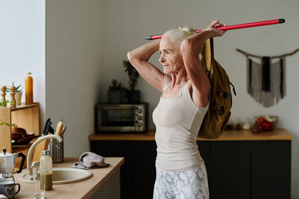 Vezető aktív nő hátizsák bottal áll a kamera előtt a konyhában, és élvezi a reggeli edzés reggeli előtt - Fotó, kép