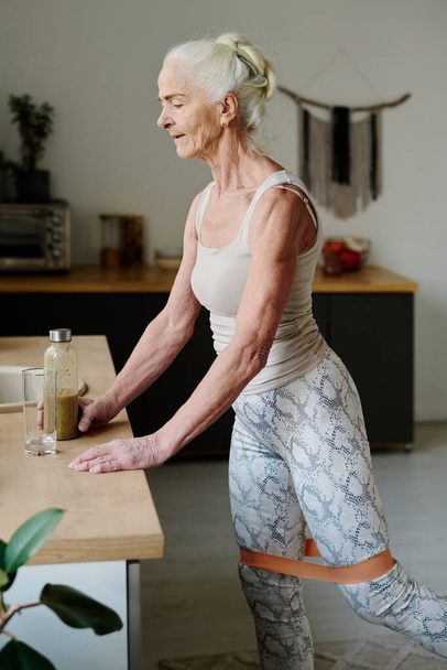 Старшая женщина в активной одежде держит бутылку домашнего смузи, стоя у кухонного стола и делая физические упражнения - Фото, изображение