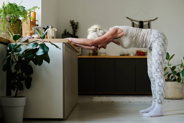 Vista lateral de la mujer jubilada activa inclinándose hacia adelante contra el mostrador de la cocina mientras está de pie en el suelo durante el entrenamiento de la mañana - Foto, imagen