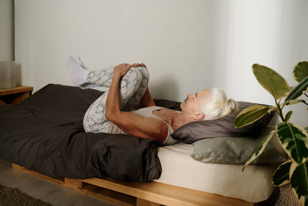 Vue latérale de la femme âgée en vêtements de sport gardant les jambes pliées dans les genoux contre sa poitrine tout en étant couchée sur le lit après le sommeil et faire des exercices - Photo, image