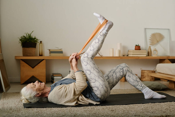Πλευρική άποψη της ηλικιωμένης ενεργού γυναίκας εξάσκηση ασκήσεις με ζώνη αντίστασης, ενώ βρίσκεται στο χαλί στο υπνοδωμάτιο και την αύξηση του δεξιού ποδιού κατά τη διάρκεια προπόνηση - Φωτογραφία, εικόνα