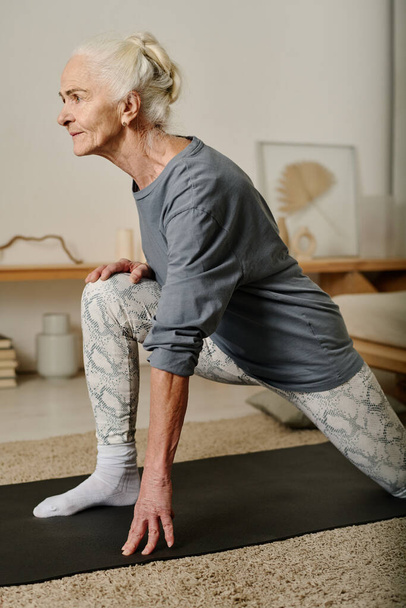 Side view of mature active woman looking forwards while standing on mat with one leg bend in knee and the other one stretched backwards - Photo, Image