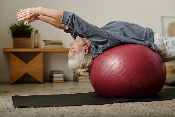 Seitenansicht einer grauhaarigen Frau in Aktivkleidung, die auf einem Fitball liegt und die Arme nach hinten streckt, während sie körperliche Übungen auf Matte macht - Foto, Bild