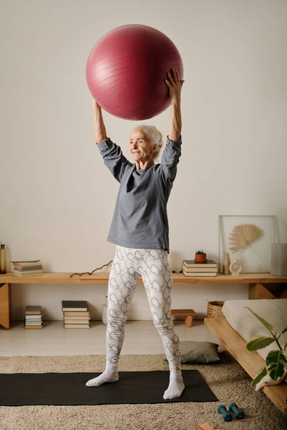 Actieve gepensioneerde vrouw in sportkleding die fitbal over haar hoofd houdt terwijl ze op de vloer van de slaapkamer staat tijdens de ochtendtraining - Foto, afbeelding
