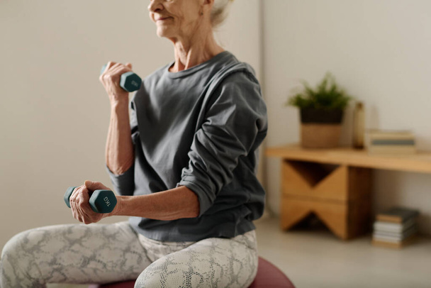 Nyugdíjas nő szürke pulóverben, súlyzókkal a kezében, miközben a kamera előtt ül, és alvás után gyakorolja a karizmok gyakorlását. - Fotó, kép