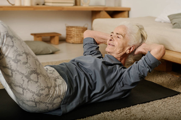 Mulher ativa sênior em leggins e pulôver de algodão cinza fazendo sit ups no tapete preto durante o treino matinal no chão do quarto - Foto, Imagem