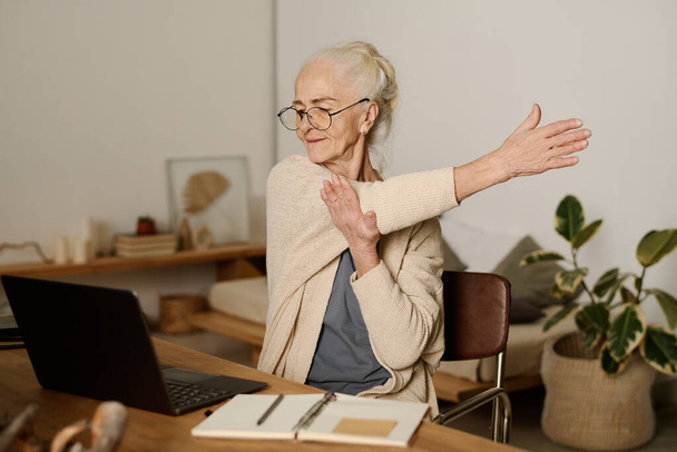 Mujer mayor en gafas de vista mirando la pantalla de la computadora portátil y haciendo ejercicio para estirarse mientras está sentada junto a la mesa y trabajando con datos en línea - Foto, Imagen