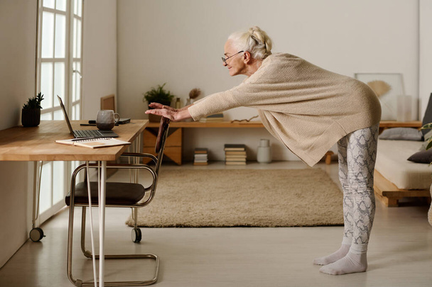 Leeftijd vrouw in casualwear kijken naar laptop scherm terwijl het vasthouden van door stoel, voorover buigen en het doen van stretching oefening - Foto, afbeelding