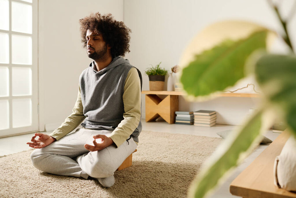 Спокойный парень в толстовке и трениках расслабляется на полу спальни, сидя в позе лотоса и медитируя по утрам - Фото, изображение