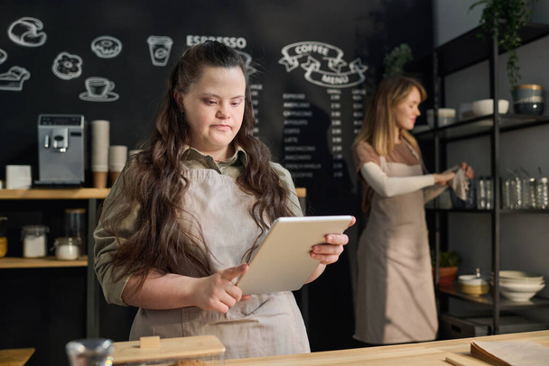 Young waitress with Down syndrome looking at tablet screen while standing by counter in front of camera against colleague - Photo, Image