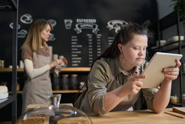 Jonge serieuze vrouw met een handicap op zoek door online menu van cafe tijdens het buigen tegen de toonbank met collega op de achtergrond - Foto, afbeelding
