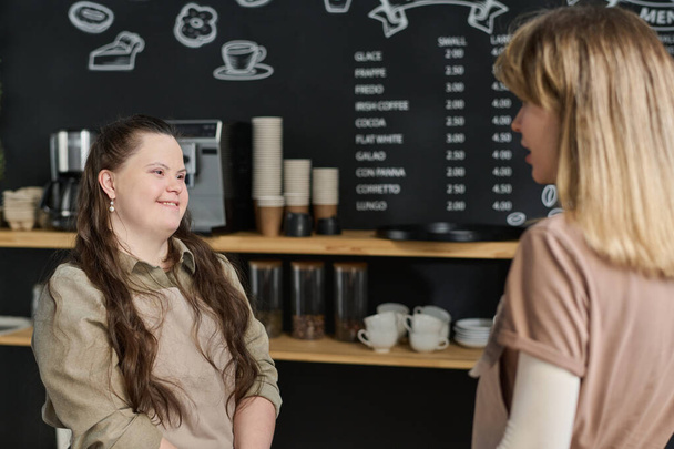 Happy young waitress with Down syndrome and her colleague having chat while standing by workplace against blackboard with menu - Photo, Image