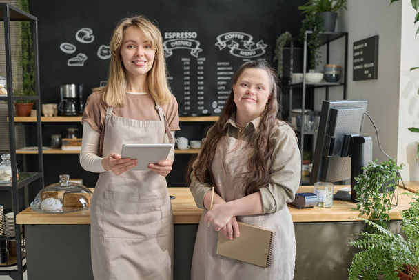 Młoda kelnerka z zespołem Downa i jej koleżanka z tabletem stojąca przy ladzie z monitorem i patrząca w kamerę - Zdjęcie, obraz