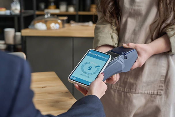 Manos de la joven camarera sosteniendo terminal de pago, mientras que las mujeres invitadas de café con smartphone con datos de operación de banca en línea en la pantalla - Foto, imagen