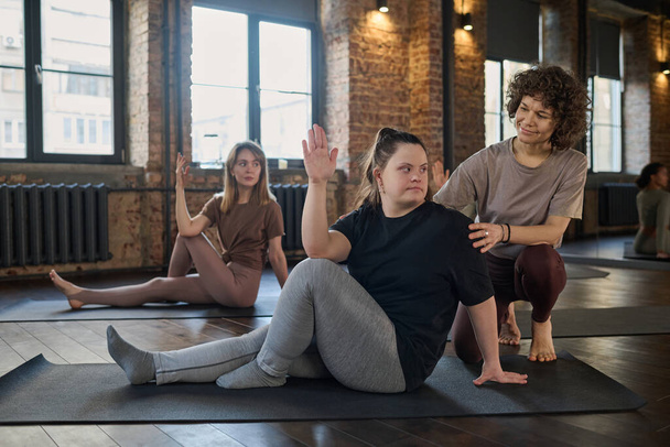 Treinador de fitness feminino jovem ajudando mulher com síndrome de Down fazendo exercício físico no tapete no ginásio contra esportista loira - Foto, Imagem