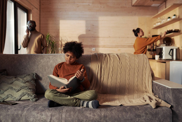 Diligente ragazzo afroamericano seduto su comodo divano grigio con cuscino e lana beige a quadri lavorati a maglia e libro di racconti - Foto, immagini
