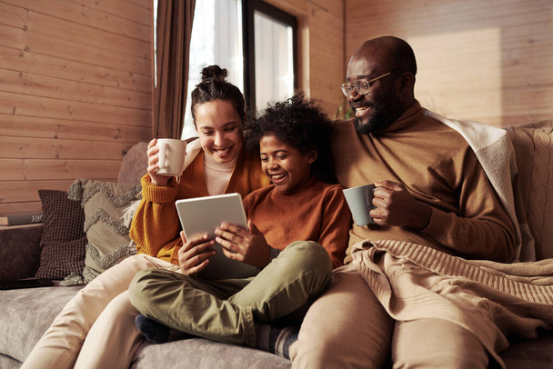 Счастливая молодая межкультурная семья из трех человек смотрит на экран планшета, который держит милый мальчик, сидя на диване и смотря ситком на досуге - Фото, изображение
