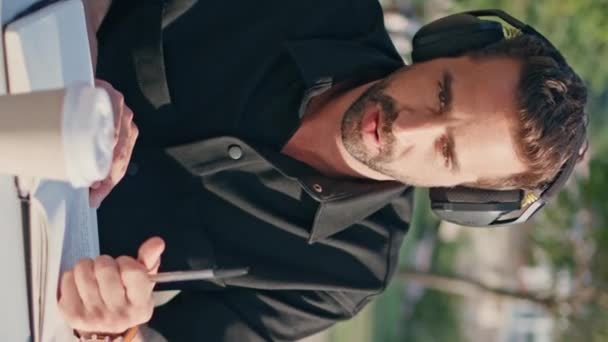 Приглушенный фрилансер слушает наушники в зеленом парке вертикального видео. Латиноамериканец держит ручку, работающую на открытом воздухе. Бородатый стратупер наслаждается музыкой в поисках вдохновения на природе  - Кадры, видео
