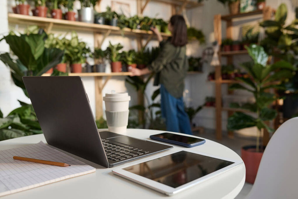 Електронні гаджети на столі в магазині рослин, робочий перевіряє заводи на полицях на фоні - Фото, зображення