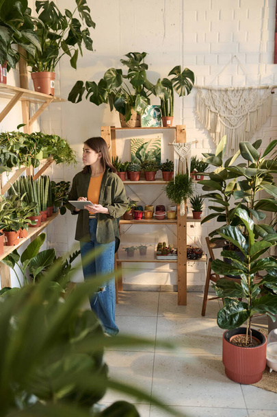 Jovem funcionário da loja de plantas verificando plantas em vasos em prateleiras com tablet em suas mãos - Foto, Imagem