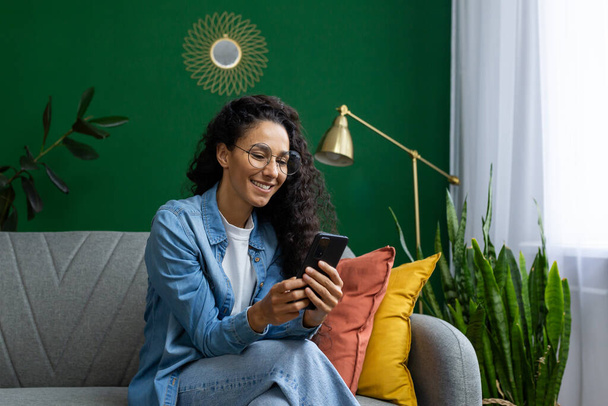 Hermosa mujer hispana en casa en el sofá en la sala de estar, la mujer sonríe contenta sostiene el teléfono en sus manos, utiliza aplicaciones en el teléfono inteligente, navega por Internet y las redes sociales. - Foto, Imagen