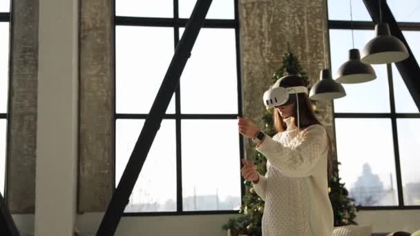 Egy elbűvölő fiatal lány virtuális valóság fejfedőt vesz fel egy karácsonyfával a háta mögött. Kiváló minőségű 4k felvételek - Felvétel, videó