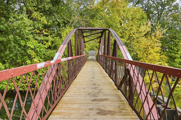 Ponte pedestre de treliça de aço vermelho rústico cercada por folhagem no início do outono em Lawton Park, Fort Wayne, Indiana, convidando a exploração serena. - Foto, Imagem