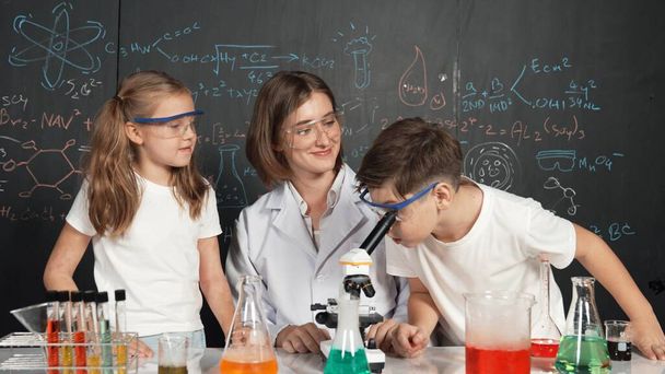 Caucasian boy looking under microscope while teacher giving advice. Professional instructor wearing lab suit looking for diverse student at table with beaker filled with colored solution. Erudition. - Photo, Image