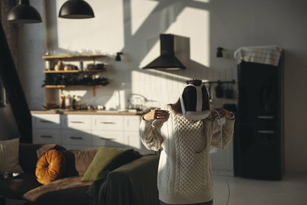 No conforto de seu apartamento, uma jovem deslumbrante joga um jogo online com um fone de ouvido VR. Foto de alta qualidade - Foto, Imagem