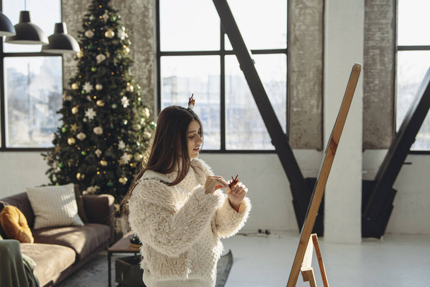 Rayonnant de bonheur, une jeune femme vibrante portant un masque de renne de Noël rayonne dans l'appartement. Photo de haute qualité - Photo, image