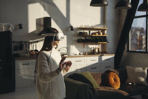 No conforto de seu apartamento, uma jovem deslumbrante joga um jogo online com um fone de ouvido VR. Foto de alta qualidade - Foto, Imagem