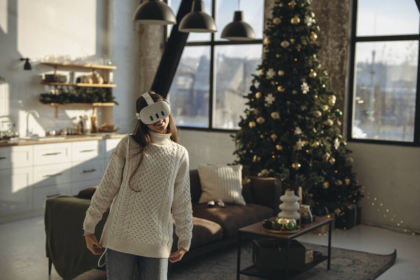 Uma jovem, imersa em um jogo online, usa um fone de ouvido de realidade virtual em casa. Foto de alta qualidade - Foto, Imagem