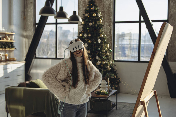 Egy karácsonyfával körülvett gyönyörű fiatal nő virtuális valóság headsetet használ. Kiváló minőségű fénykép - Fotó, kép