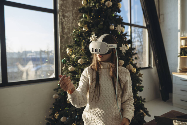 Ένα σαγηνευτικό πορτραίτο ενός κοριτσιού στολισμένο με ένα σετ κεφαλής εικονικής πραγματικότητας, πλαισιωμένο από ένα χριστουγεννιάτικο δέντρο. Υψηλής ποιότητας φωτογραφία - Φωτογραφία, εικόνα