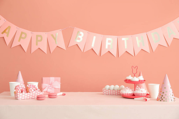 Süßigkeiten auf weißem Tisch und Papiergirlanden zum Geburtstag an dunkelbeiger Wand - Foto, Bild