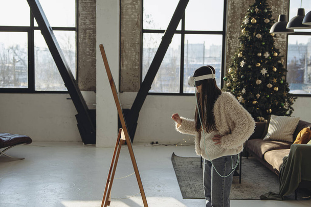 Con el ambiente navideño imperante, una mujer joven con atuendo ligero y un auricular VR se mira al espejo. Foto de alta calidad - Foto, imagen