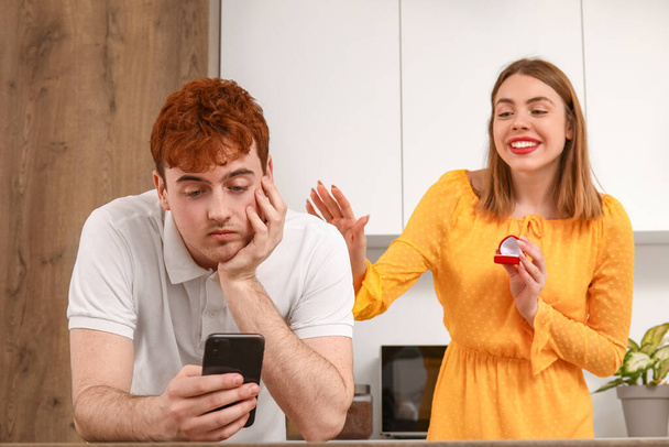 Młoda kobieta z pierścionkiem zaręczynowym i jej chłopak za pomocą telefonu komórkowego w kuchni - Zdjęcie, obraz