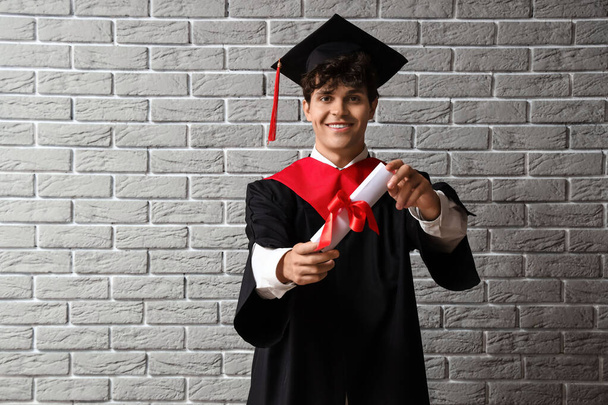 Male graduate student with diploma on grey brick background - Foto, afbeelding
