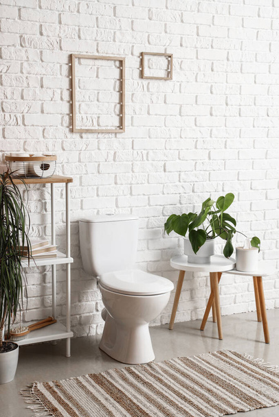 Innenraum der hellen Toilette mit Keramik-Toilettenschüssel, Regal und Zimmerpflanze in der Nähe der weißen Ziegelwand - Foto, Bild