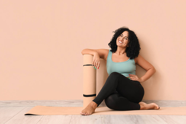 Sporty young woman with yoga mat sitting on floor against beige background - Photo, Image