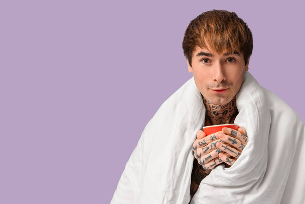 Bello giovanotto con coperta e tazza di tè su sfondo lilla, primo piano - Foto, immagini