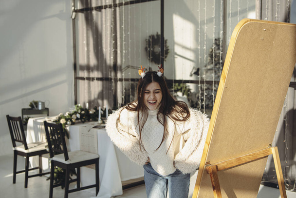 Com a luz do sol inundando, uma jovem mulher vibrante desfruta de uma máscara de rena de Natal em seu apartamento. Foto de alta qualidade - Foto, Imagem