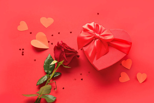 Hermosa flor de rosa, caja de regalo en forma de corazón y corazones de papel sobre fondo rojo. Celebración de San Valentín - Foto, Imagen