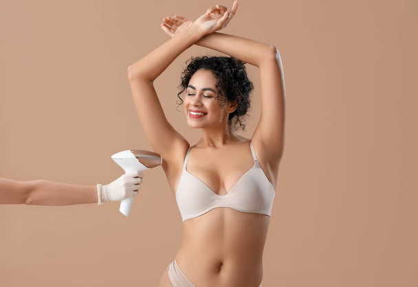 Krásná žena ve spodním prádle a ruce kosmetičky s fotoepilátorem na béžovém pozadí - Fotografie, Obrázek