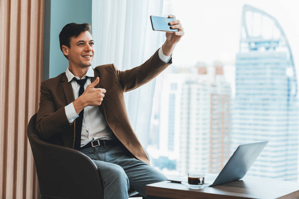 Επαγγελματίας επιχειρηματίας βγάζει selfie κοντά στο παράθυρο με ουρανοξύστη ενώ δίνει τον αντίχειρα στην κάμερα. Εξειδικευμένος διευθυντής φωτογραφίζει ενώ κάθεται στο τραπέζι με την κούπα του καφέ και το φορητό υπολογιστή. Διακοσμημένα. - Φωτογραφία, εικόνα